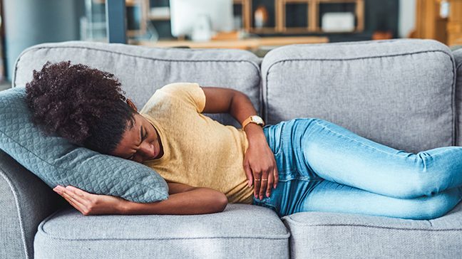 Young, slender black woman in jeans and yellow sweater lying on gray couch holding abdomen with furrowed brow