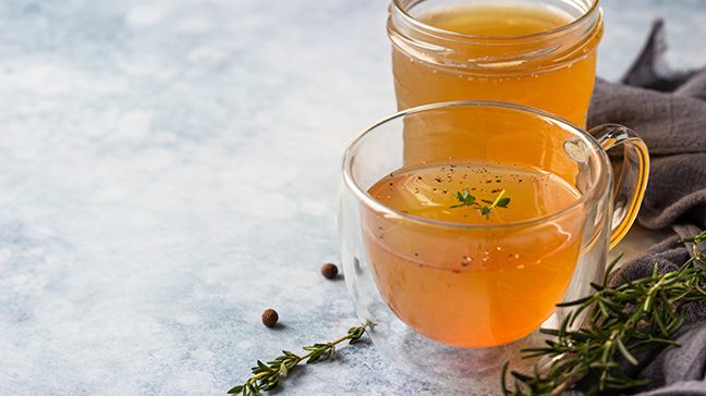 Bone broth in a clear mug and a clear mason jar on a marble countertop