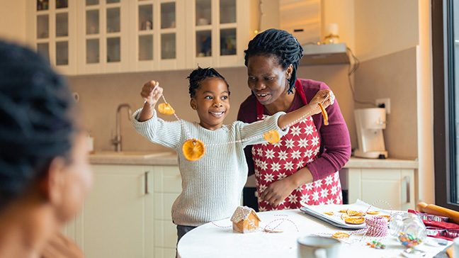 A young girl in a grey sweater holds a garland made of dried oranges while an adult woman in a red apron looks on.