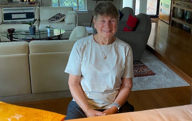 Andrea Spica smiling sitting in a chair.  A beige sofa and glass coffee table with plants on it sit behind her.