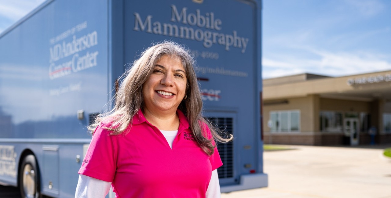 A woman with long brown hair wearing a pink polo shirt with a white long sleeve underneath smiles in front of a blue mobile mammography van