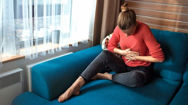 Woman on sofa holding her abdomen and showing signs of heartburn