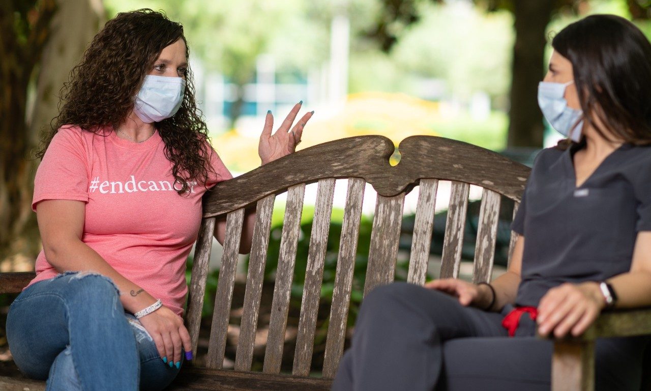 Jessica King sits on a bench outdoors with physician assistant Jill Flynn. Both women are wearing medical-grade face masks.