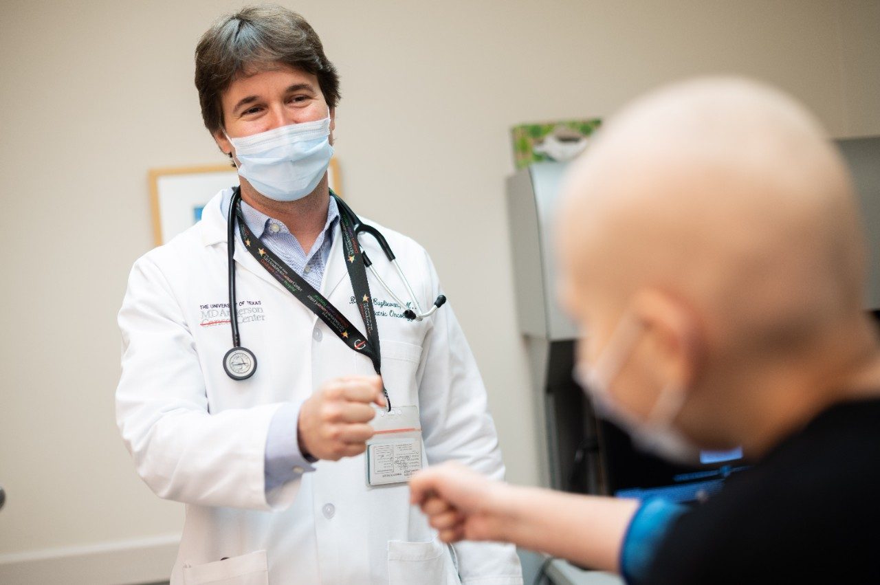 Branko Cuglievan, M.D., wearing his white coat, reaches out to fist bump with a childhood leukemia patient in the clinic while wearing his medical-grade face mask