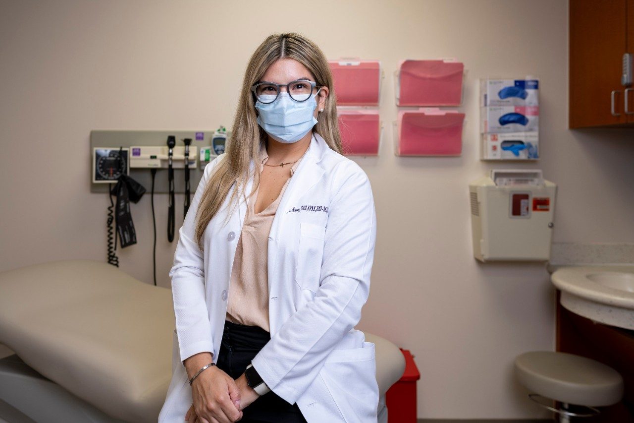 Latinx nurse Ashley Martinez sits with her hands on top of each other in a clinic room with folders hanging in the background behind her.