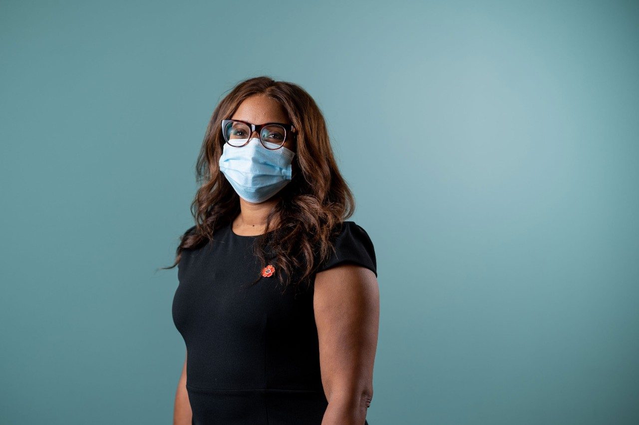 Portrait of Lorna McNeill, Ph.D., against a blue background. Image shows a Black woman with long brown hair and brown eyes wears brown-rimmed glasses, a blue surgical face mask and a black dress. 