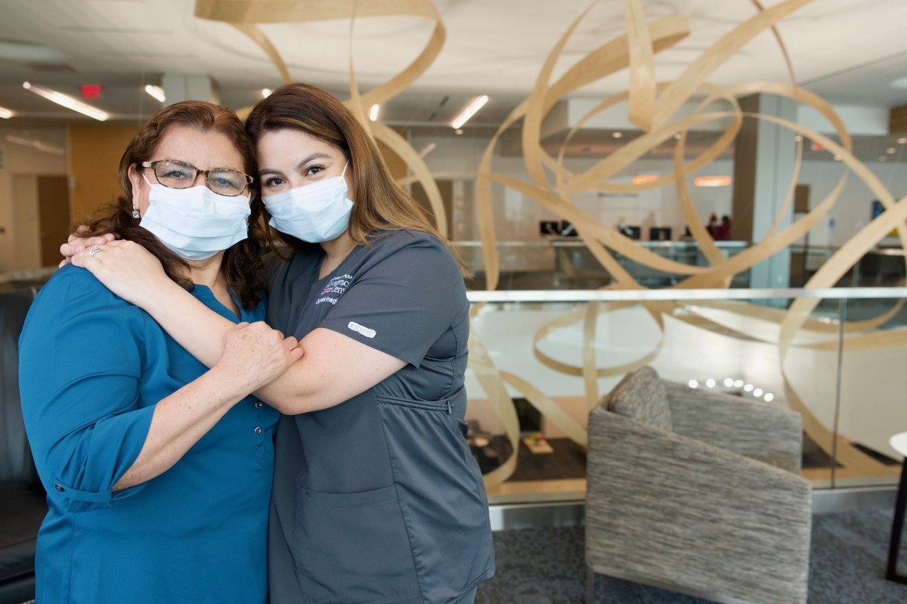 Caregiver Judy Garcia (right), stands with her arms around her mother, Maria, in the waiting area at MD Anderson The Woodlands. Judy is wearing her scrubs, and both women are wearing medical-grade face masks.