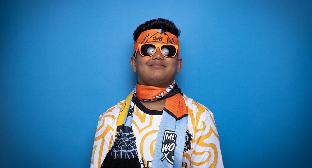 Arav Patil wearing Houston Dynamo FC gear, including orange sunglasses, a headband, a kit and a scarf, while standing against a blue background