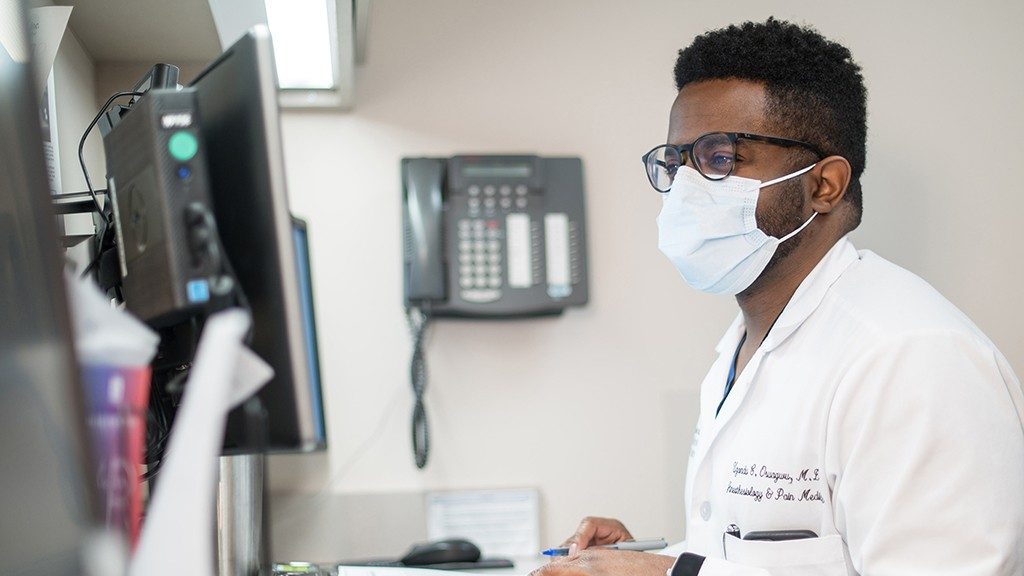 Uzondu Osuagwu, M.D., looks at a computer monitor while wearing his white coat and a medical-grade face mask