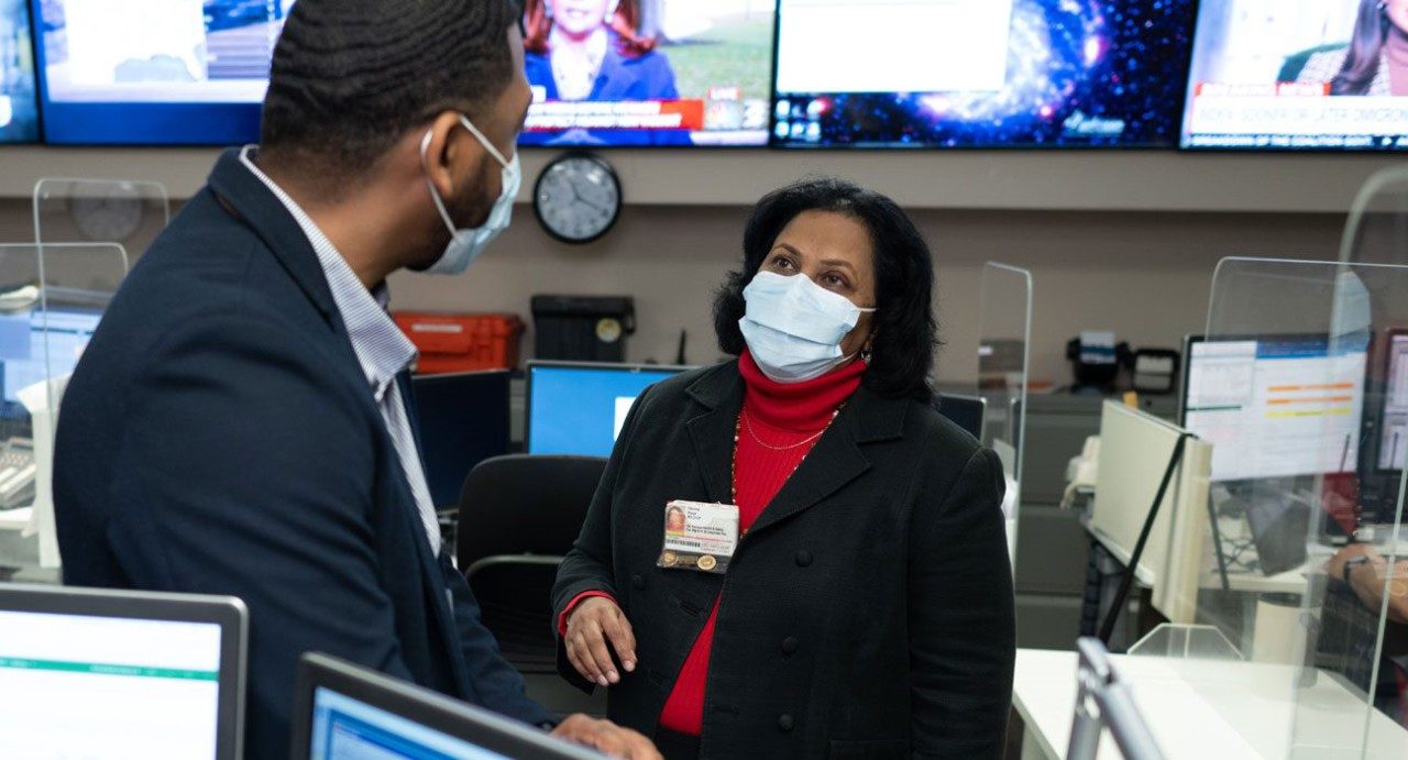 Devina Patel consults with a male colleague; TVs showing the weather forecast are in the background. Both employees are wearing medical-grade face masks.