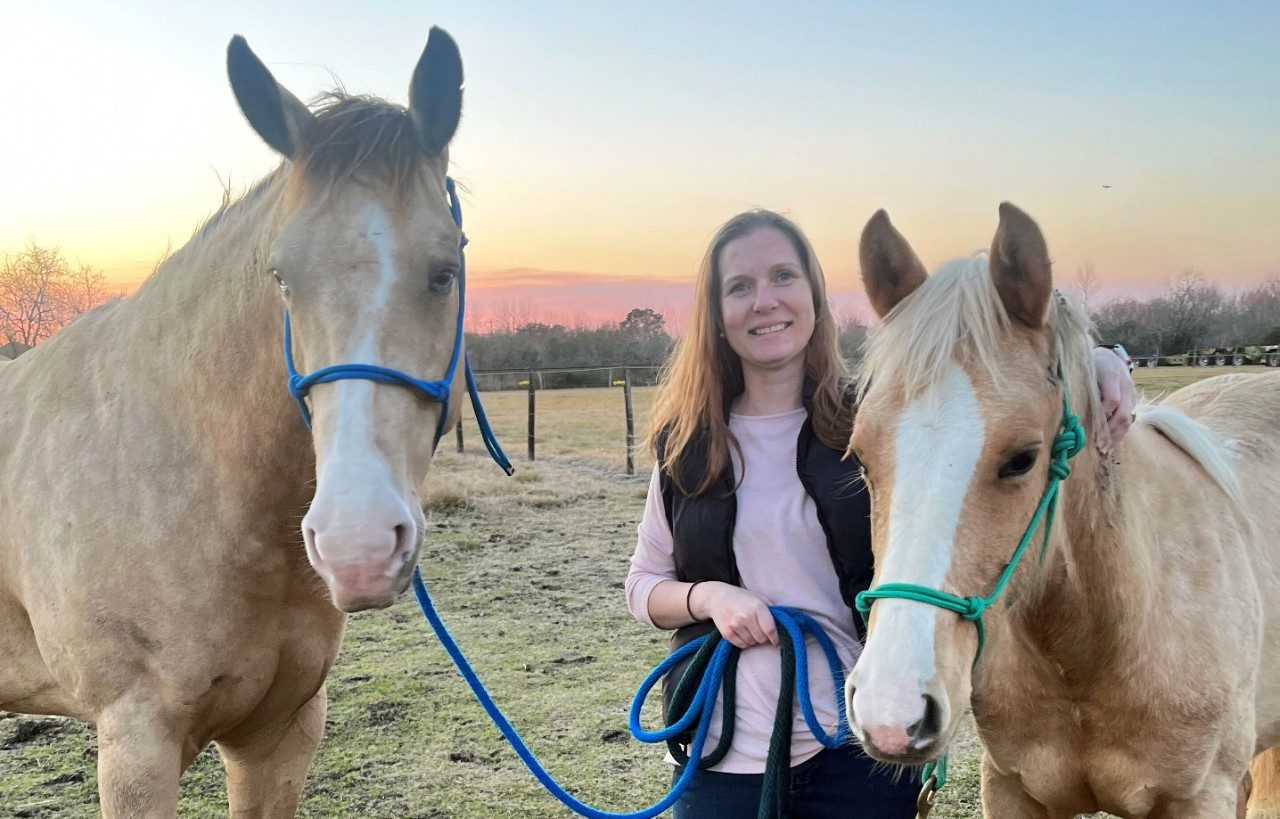 Michelle Hildebrandt, Ph.D., stands between two of her horses. She has long brown hair and is wearing jeans and a dark vest.