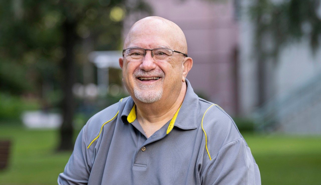 Bladder cancer patient Ron Speidel smiles as he sits outside with green grass and trees behind him. He is wearing glasses and has a goatee.