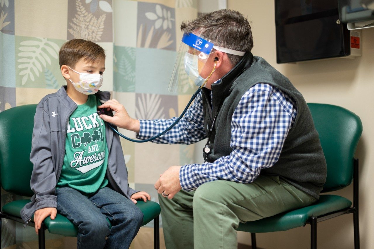 Medical oncologist Douglas Harrison, M.D., listens to the heart of pediatric rhabdomyosarcoma patient Sawyer Hack, sitting down in a clinic examination room. 