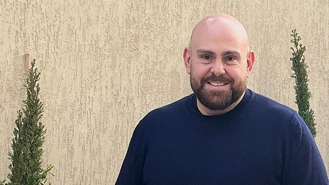 Non-Hodgkin lymphoma survivor Omar Dirani stands in front  of a wall with shrubs on either side. He has a beard and bald head and is wearing a blue shirt.