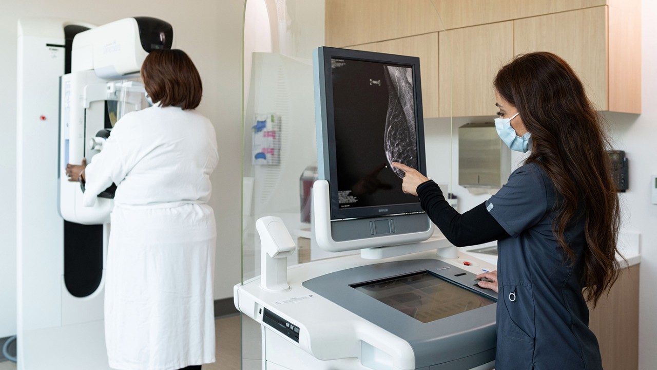 A person in a white robe faces a mammogram machine at MD Anderson the Woodlands while a Latina mammogram technician examines a digital image of a breast on a screen.