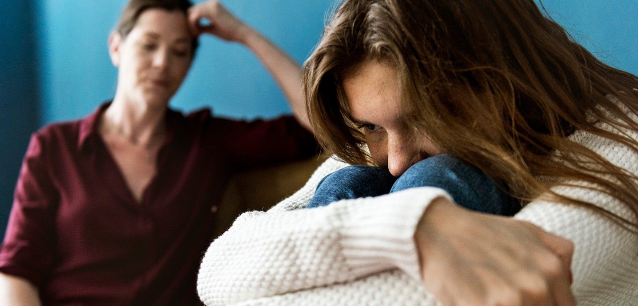 Picture of clearly distressed young person hugging her knees with equally frustrated parent sitting on couch in background with hand to her head