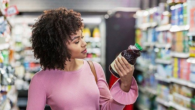 Woman with curly brown hair and a pink shirt stands in a grocery store aisle looking at the nutrition information on a bottle of purple juice