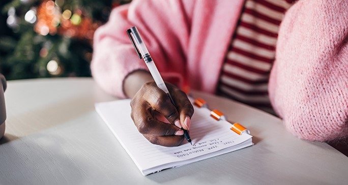 A person wearing a pink sweater and red striped shirt holds a pen and writes in a notebook