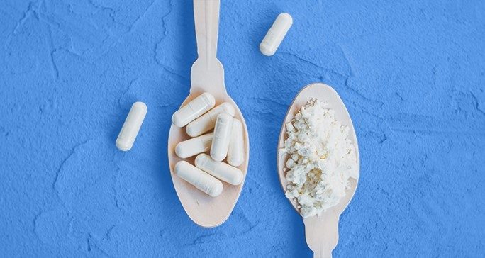 Two cream-colored spoons on a blue surface. The left spoon holds seven cream colored capsules; two additional capsules lay on each side of the spoon. The right spoon holds a white powder.