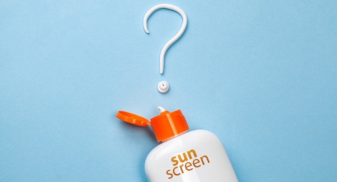 An white sunscreen bottle with an open orange cap and text lies underneath a white question mark made of sunscreen.