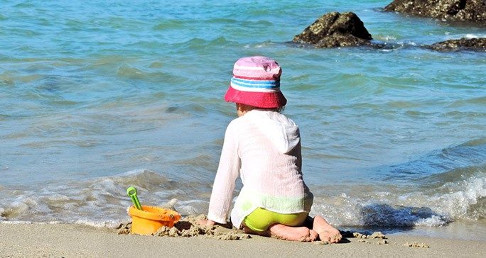 Rear view of young child wearing a red bucket hat, white long-sleeve shirt and lime green swim suit playing in the sand at the beach