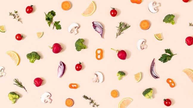 Food including cherry tomatoes, peppers, radishes, carrot slices, mushrooms, orange slices, broccoli, onion slices and herbs are laid out on a pink surface.