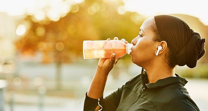 A woman wearing an athletic jacket, headphones and a hair wrap drinks from a clear bottle.