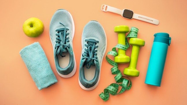 A blue workout towel, blue water bottle, blue running shoes, pink fitness tracker, green apple and a green measuring tape wrapped around green dumbbells sit on an orange surface 