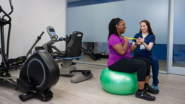 A woman in blue scrubs sits next to a women wearing workout gear holding a yellow resistance band and sitting on a green exercise ball.