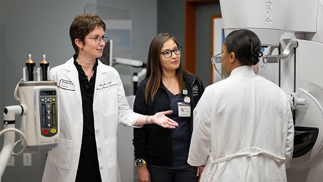 A physician in a white coat stands next to a mammography machine. Beside her, there is a woman in navy scrubs. A woman wearing a white robe stands with her back to the camera.