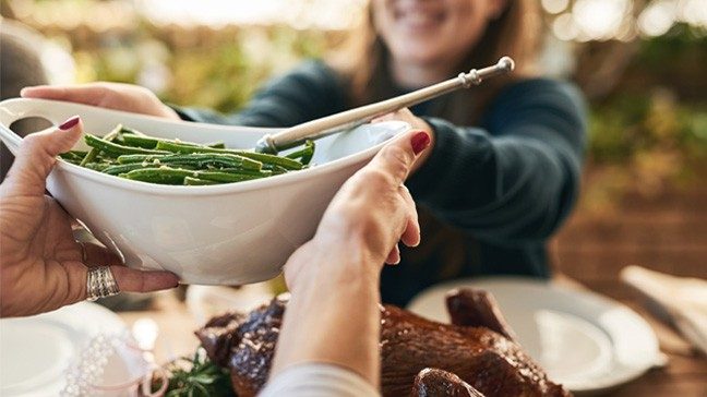 Two people at a table pass a white dish with green beans