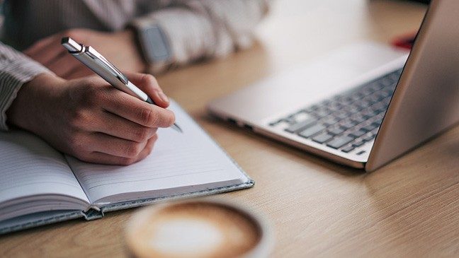 Person sitting at a table with a laptop and latte writes in a notebook with a pen