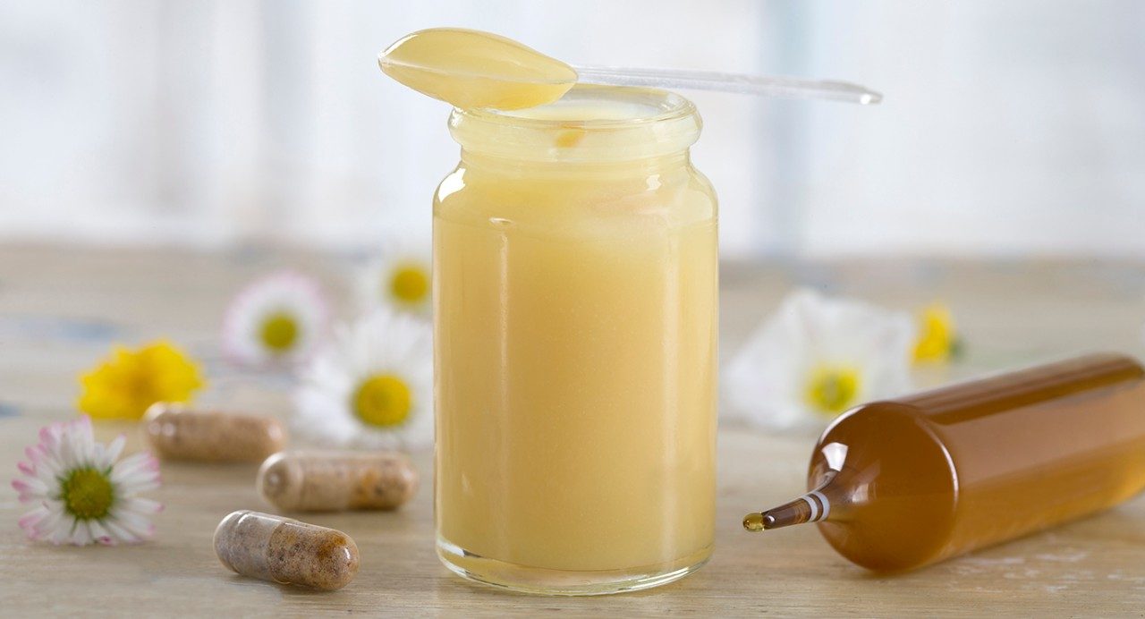 A jar of yellow royal jelly with a spoon resting on top sits on a counter alongside three supplement capsules, white flowers and a tube of royal jelly.