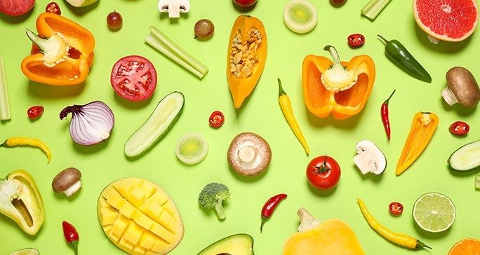 Whole and sliced fruits and vegetables on a lime green background including onion, grapes, broccoli, cucumber, tomatoes, mushroom, squash, orange peppers, chili peppers, limes, mango, zucchini and grapefruit.